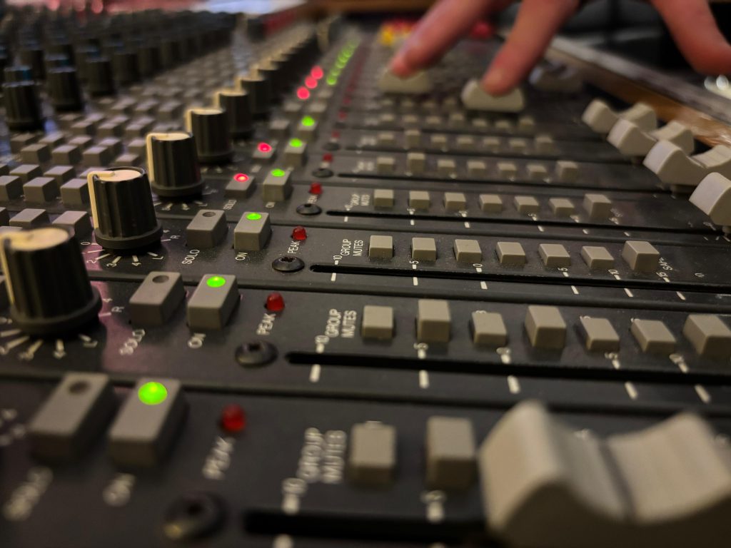 Nahaufnahme eines professionellen Audio-Mischpults mit Fadern, Knöpfen und LED-Anzeigen, eine Hand justiert die Pegel.